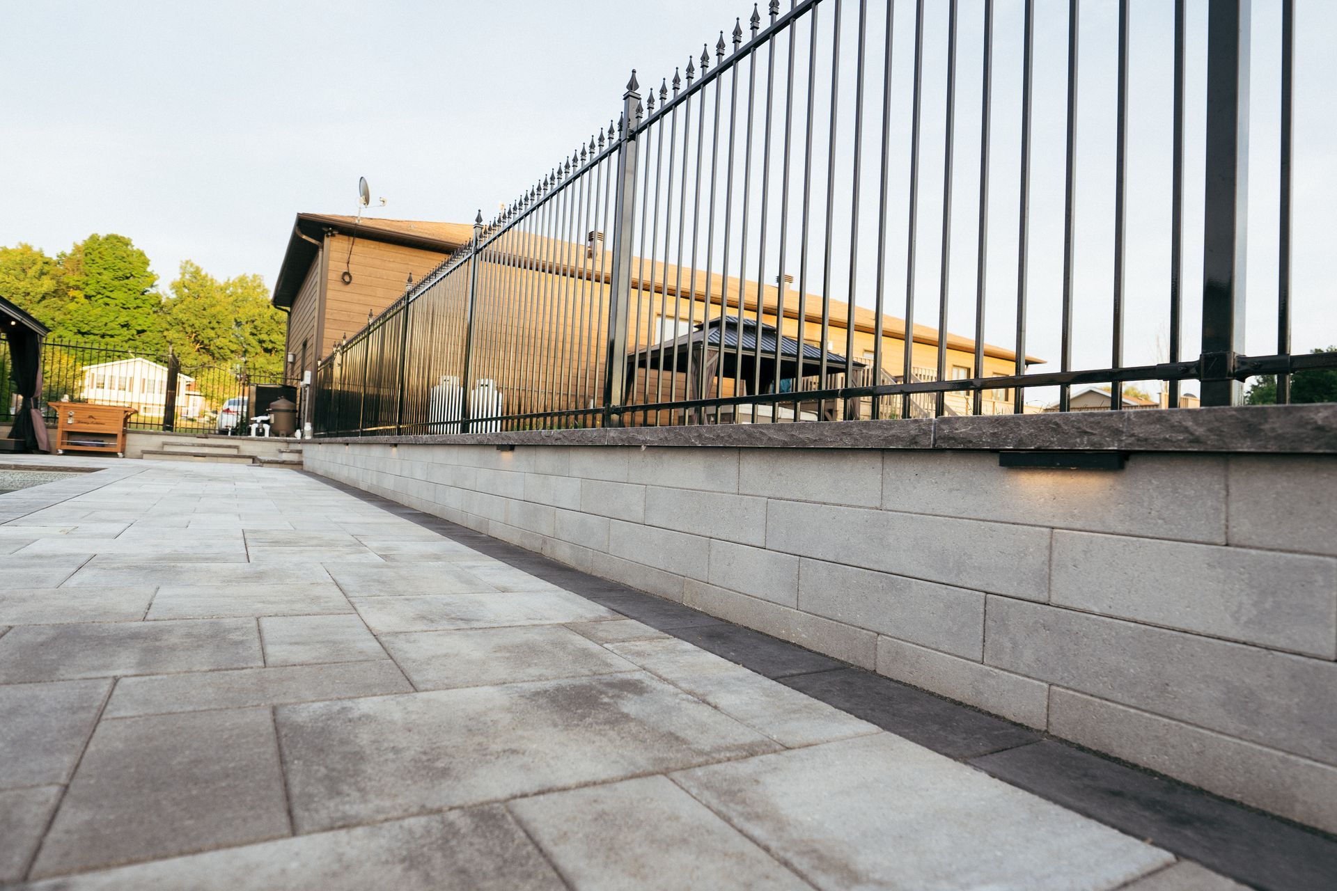 Stone walkway alongside a low retaining wall topped with a black metal fence, leading to a building.