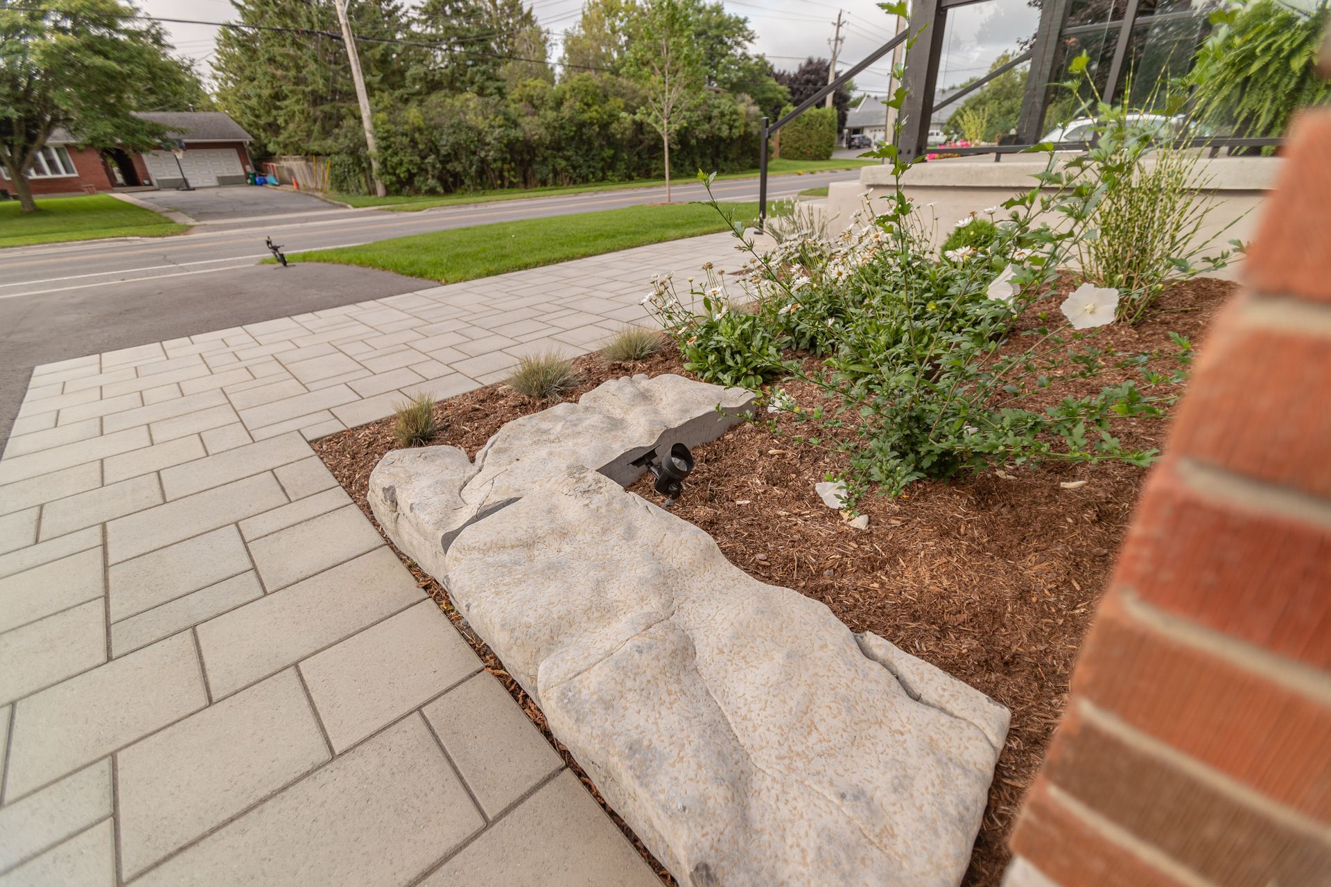 Stone planter with mulch and greenery beside a gray paved sidewalk and street.