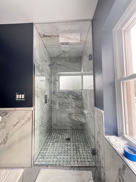 A marble-tiled shower with glass doors, next to a blue wall and a window.