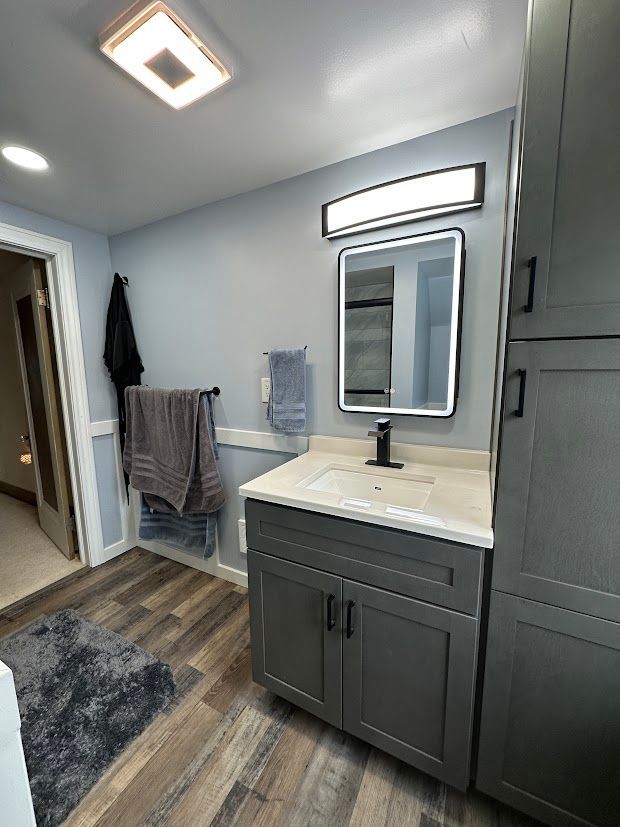 Gray bathroom with vanity, mirror, and storage cabinet, featuring light gray walls and wood-look flooring.