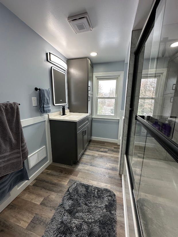 A modern bathroom with blue walls, gray vanity, and shower. Wooden floors and fluffy gray bath mat.