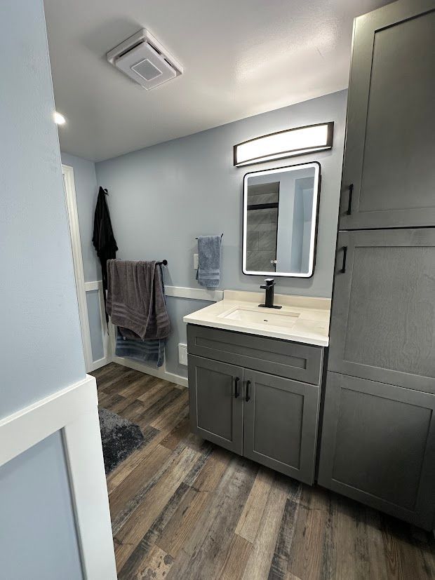 Gray bathroom with vanity, mirror, and tall cabinet. Blue walls, wood-look floor, and towels.