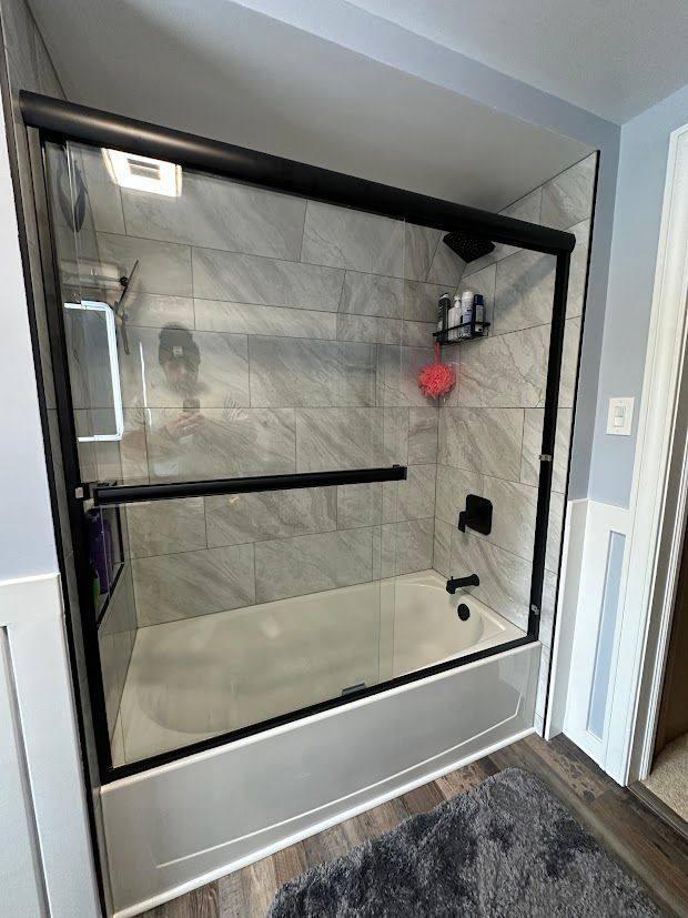 Black-framed shower door over a white tub with gray tile walls.