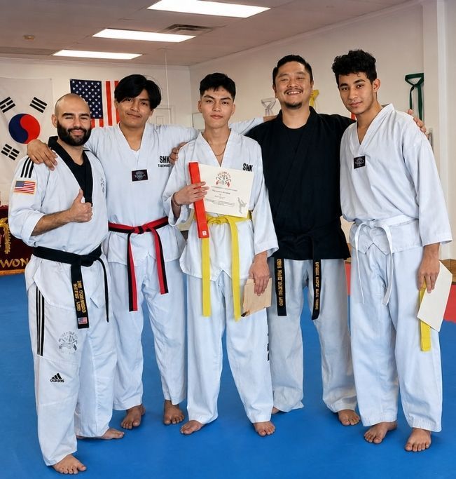 Family in Jiu-Jitsu gis poses. Indoors, blue mats, a smiling woman in the middle, around a seated man with a beard.