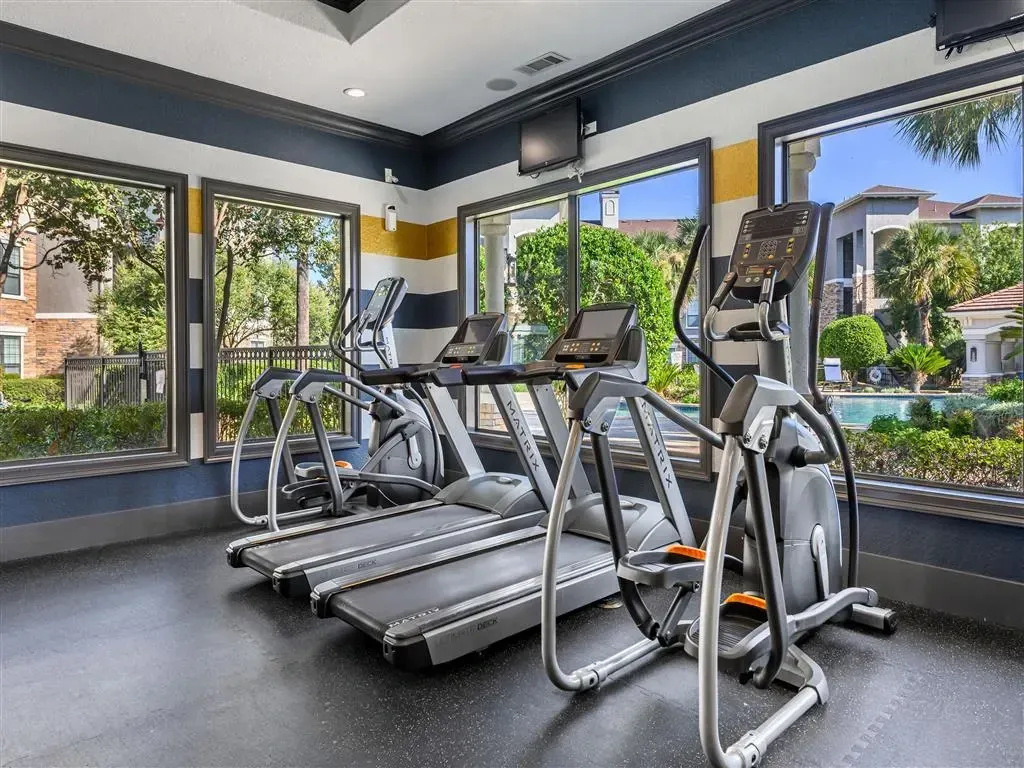 Treadmills and ellipticals in a fitness center with views of the pool and apartments.