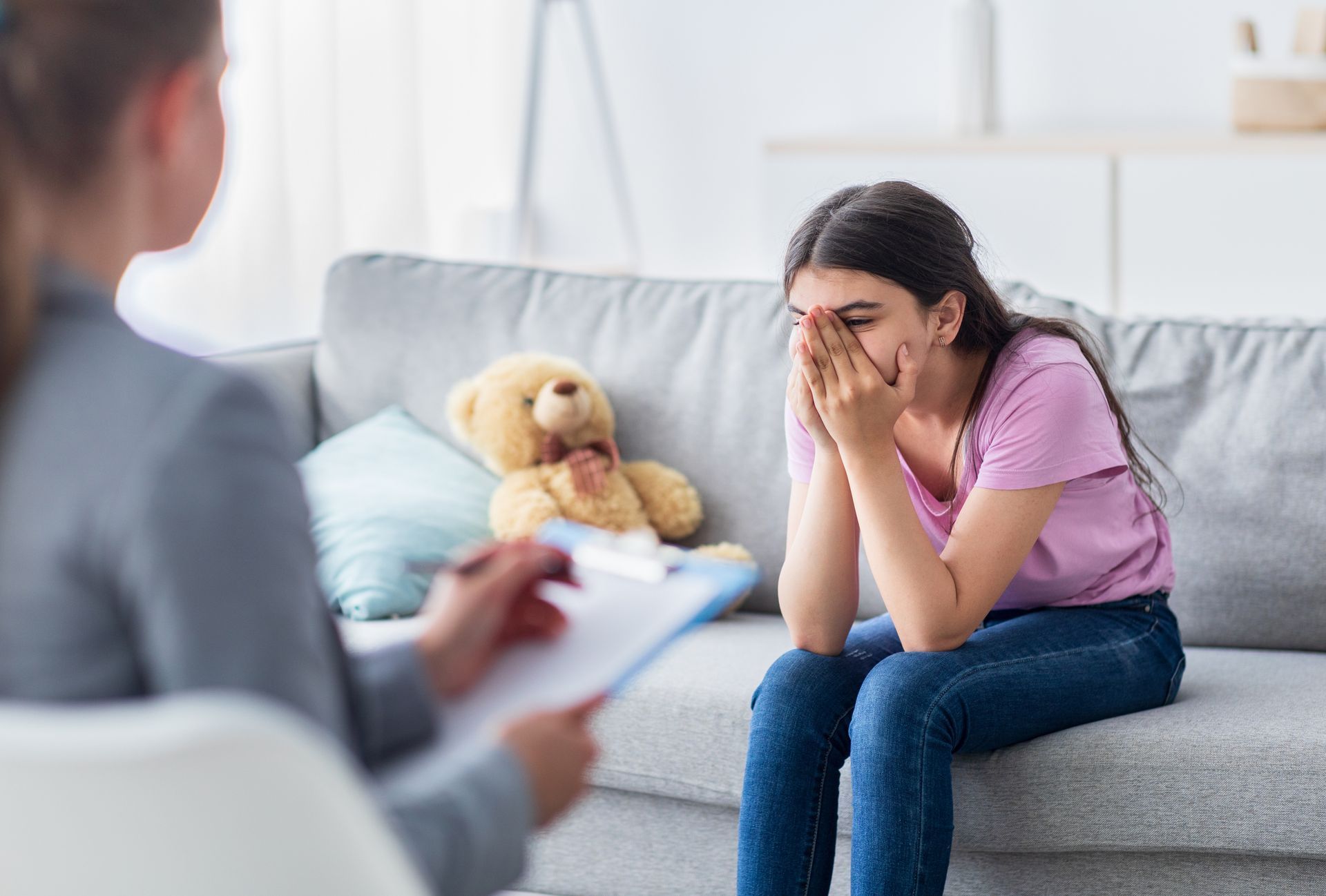 Woman sits on a couch with her face in her hands while a counselor listens nearby; teddy bear on the sofa. Woman sits on a couch with her face in her hands while a counselor listens nearby; teddy bear on the sofa.