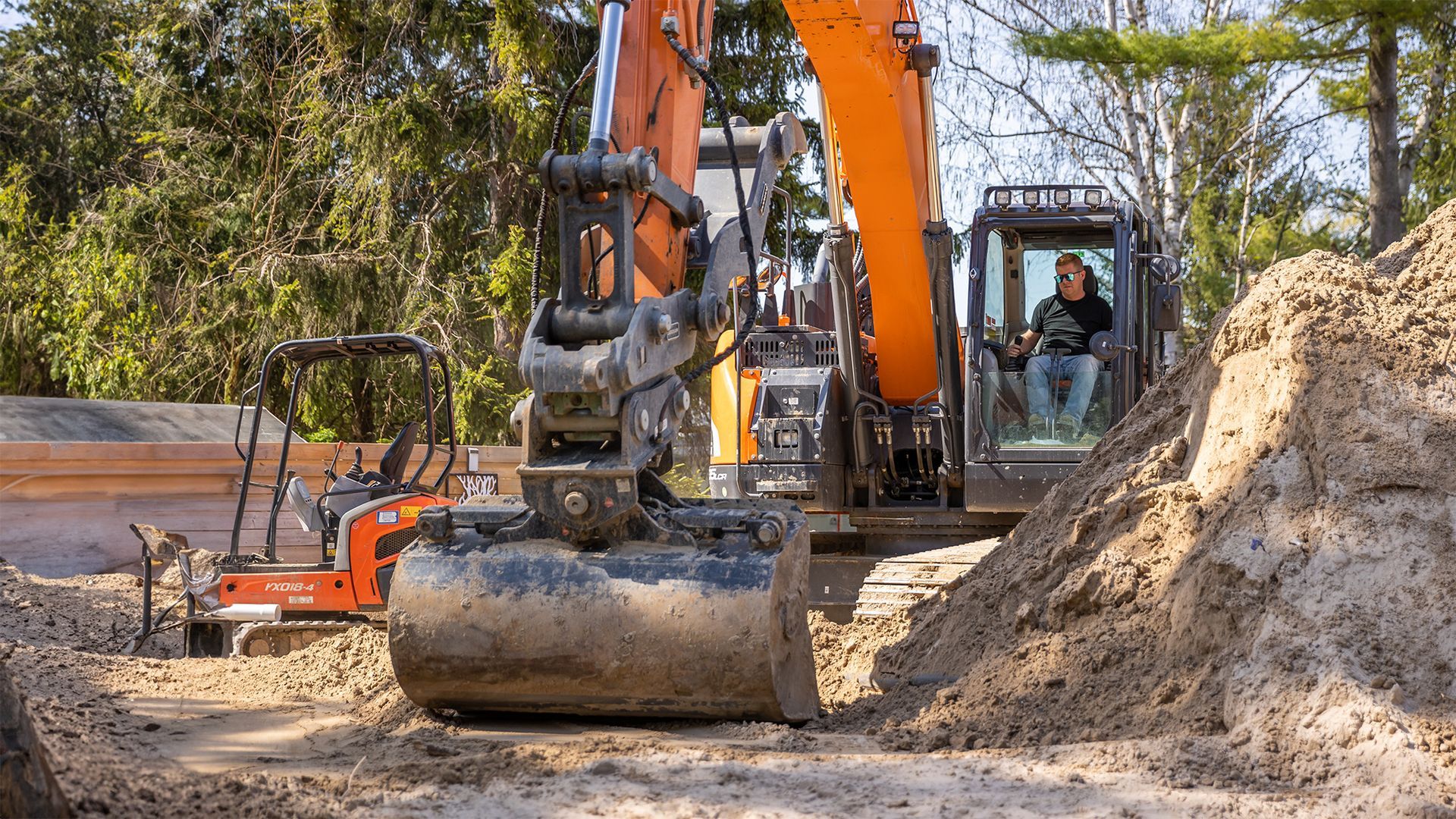 Excavator and orange compactor at a construction site with dirt piles and trees
