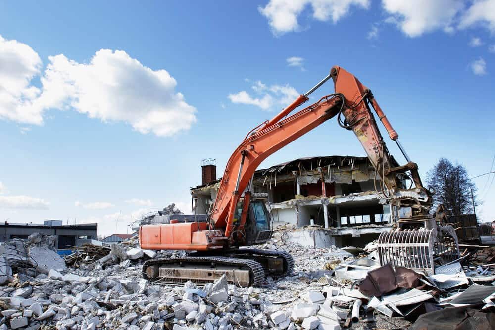 Orange excavator demolishing a building amid rubble under a blue sky