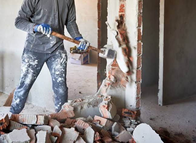 Worker using a sledgehammer to break down a concrete wall in a room with rubble on the floor