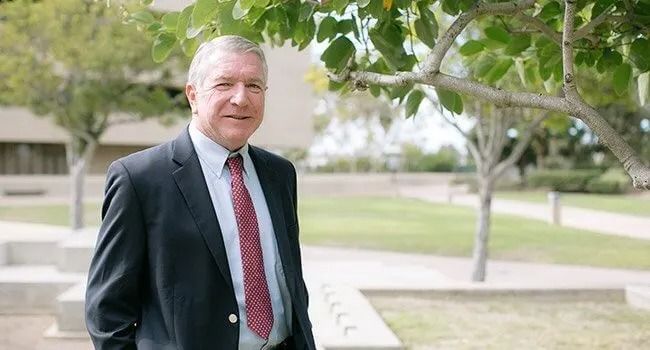 A man in a suit and tie is standing in front of a tree.