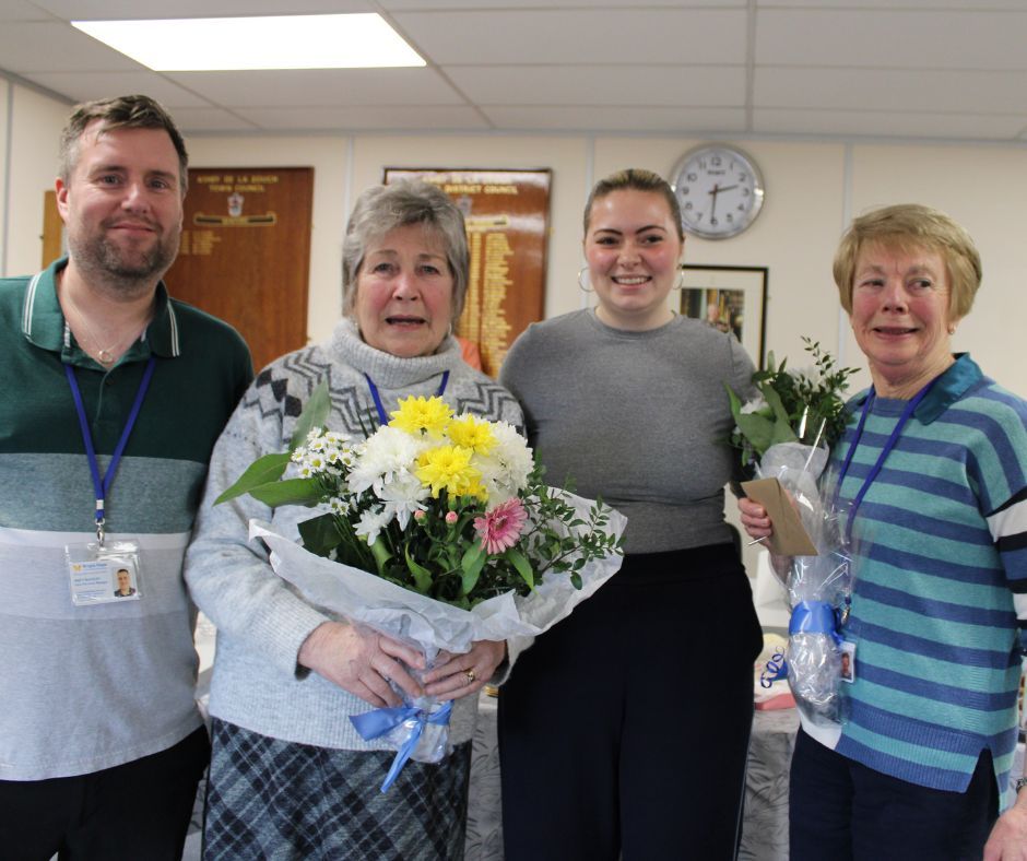 Four coworkers posing indoors, two holding bouquets and smiling in an office break room