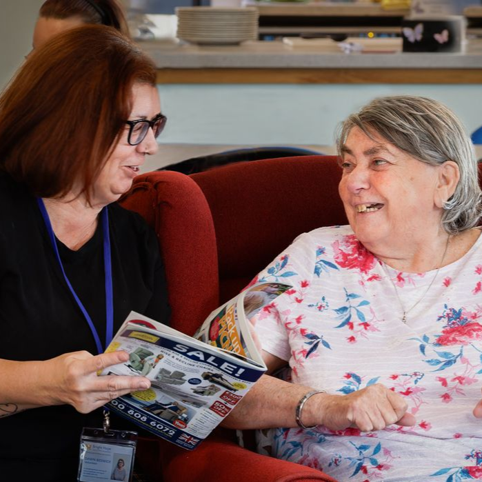 Two women smiling and reading a brochure together on a couch.