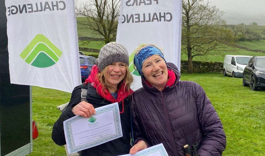 Two smiling people hold certificates outdoors beside checkered race flags and a grassy field.