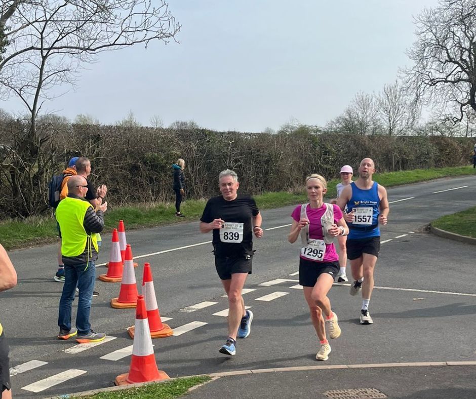 Runners pass orange cones on a road during a race, with spectators and hedges in the background.