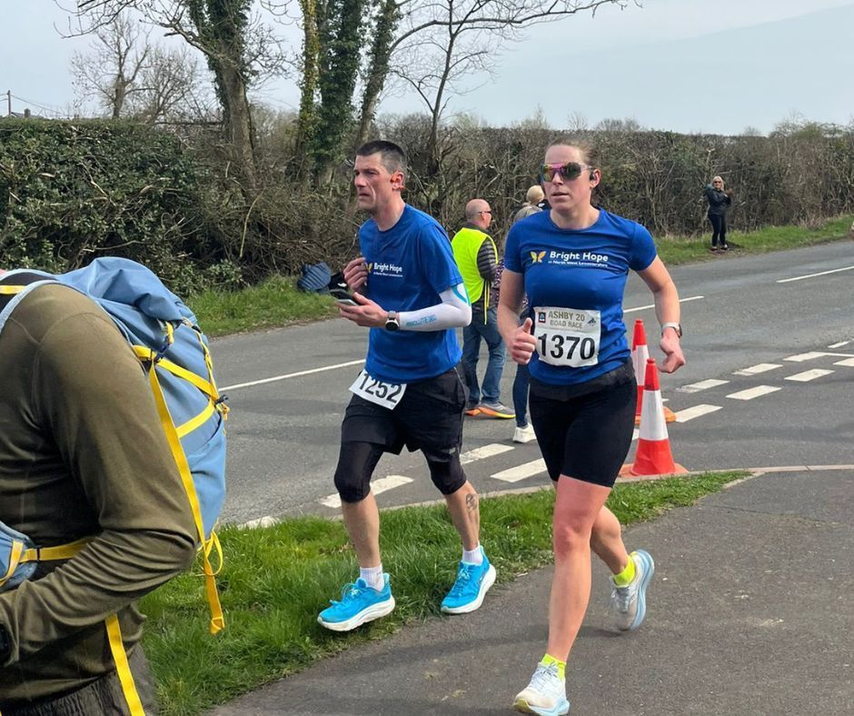 Two runners in blue shirts race on a road, one wearing bib 1370, with orange cones and hedges nearby.