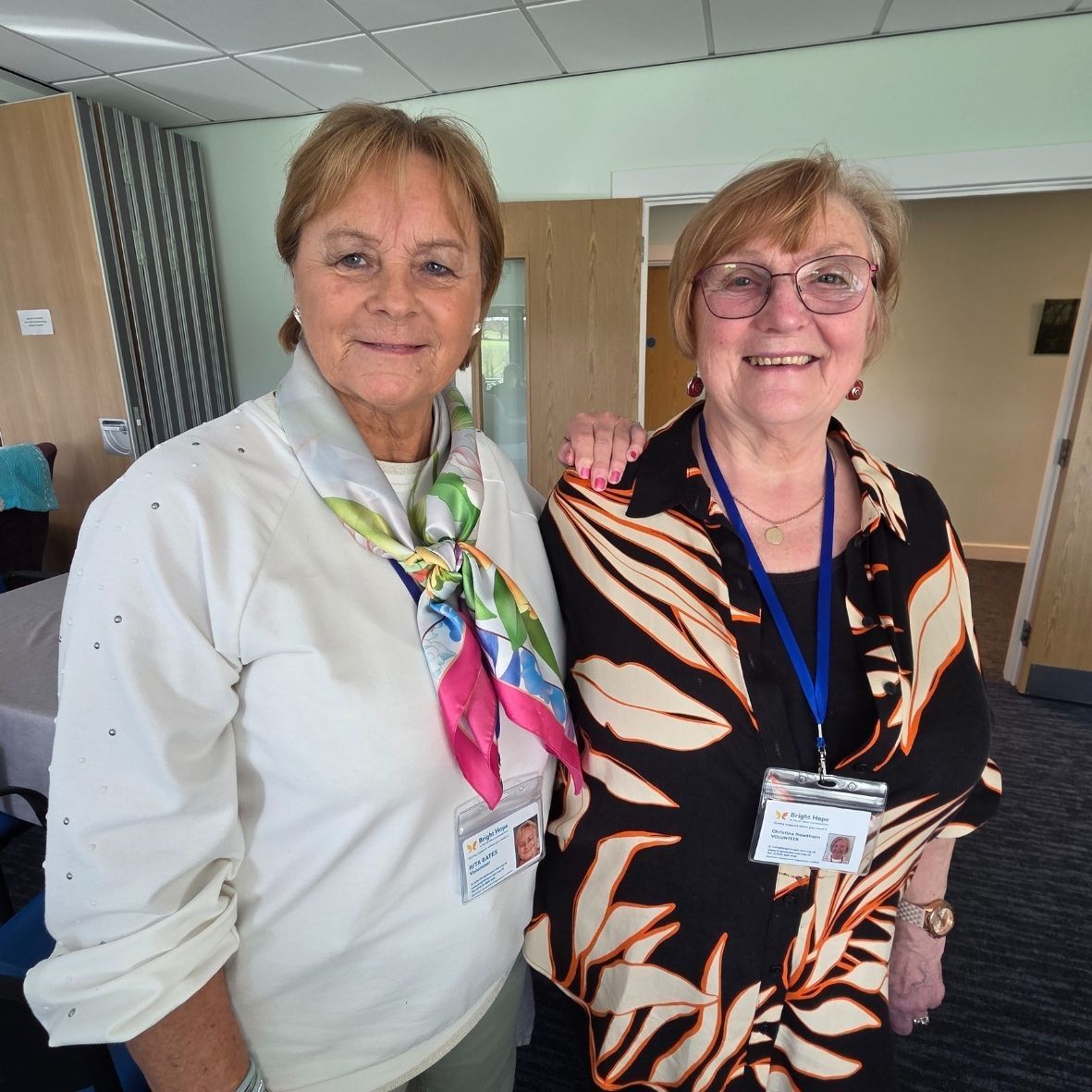 Two women smiling indoors, wearing lanyards and standing close together.