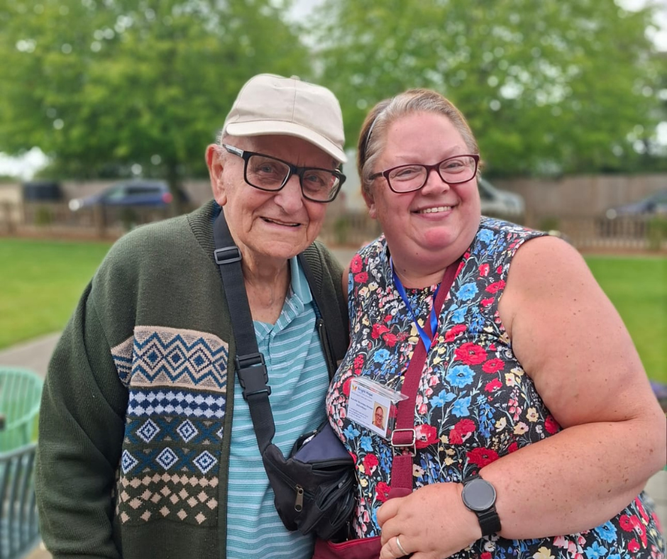 Two smiling people standing outdoors, one in a cap and sweater vest, the other in a floral top with a lanyard.