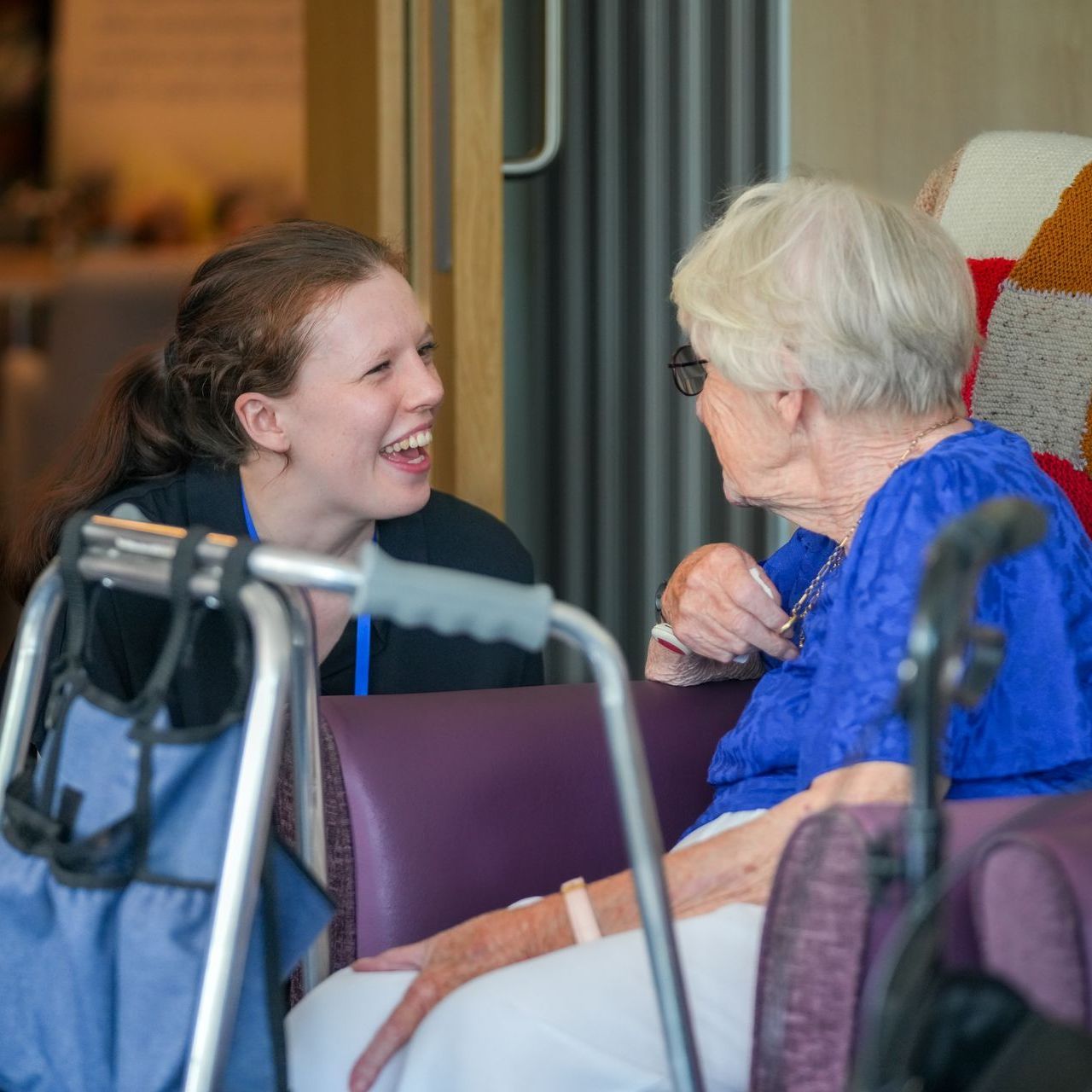Woman smiling and talking with an elderly person in a wheelchair indoors