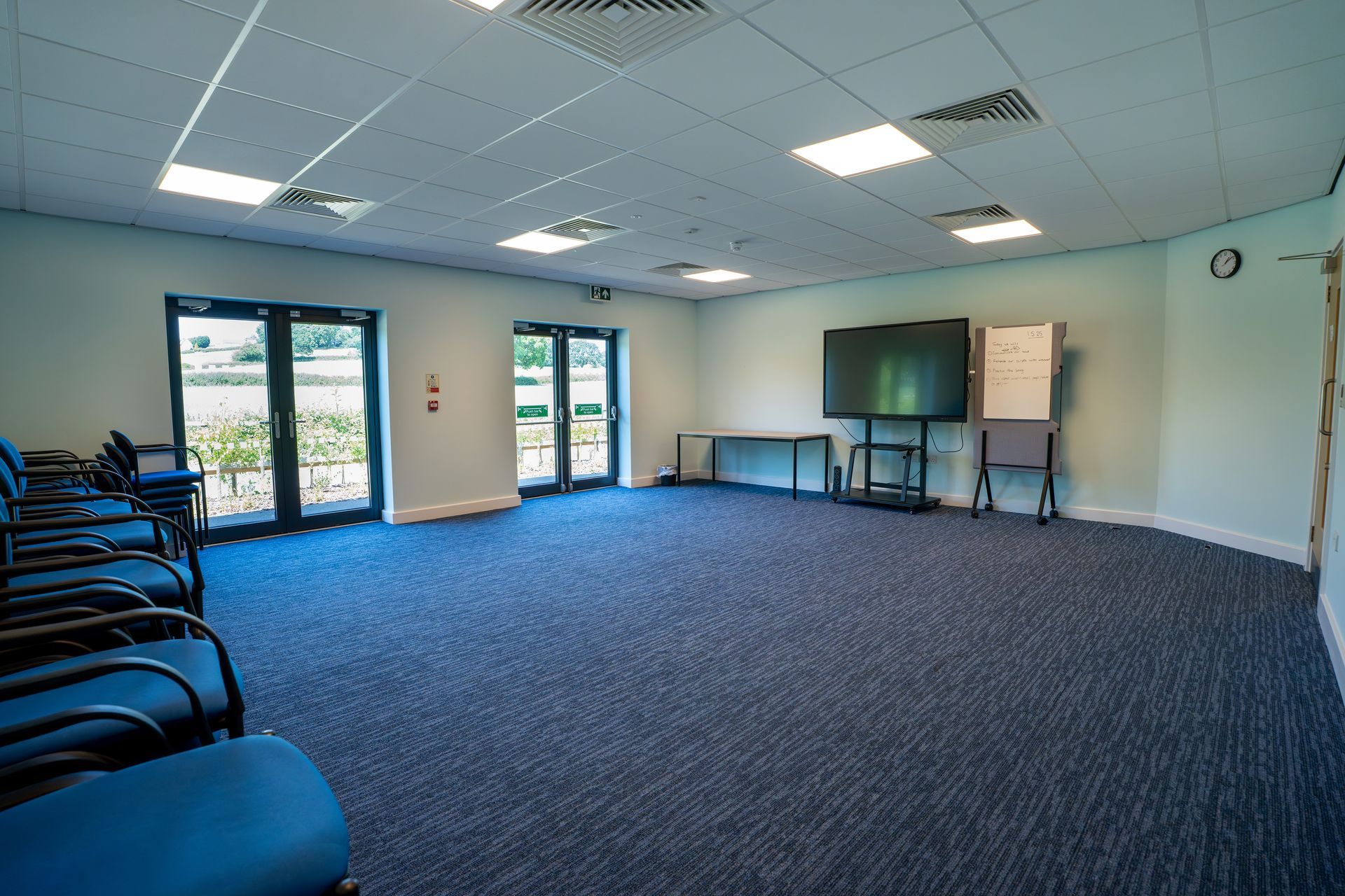 Empty meeting room with blue carpet, chairs, a TV on a stand, and large windows on the left.