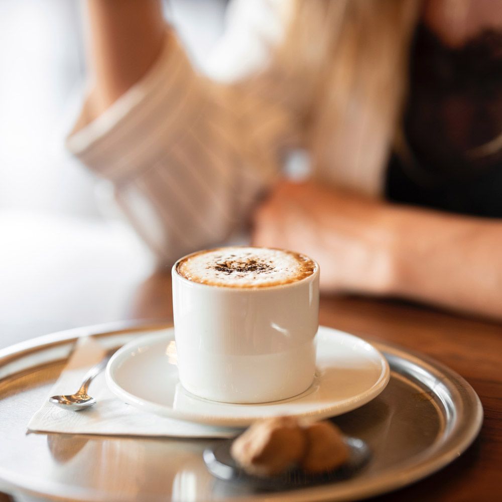 White cappuccino in a cup on a saucer with a spoon and pastry on a tray, softly blurred café background