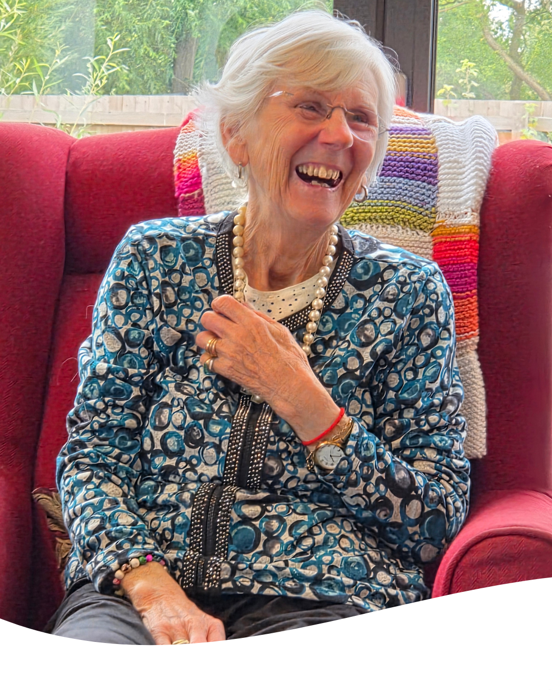 Smiling woman in a blue patterned top sits on a red chair, hand on chest, in a bright room.