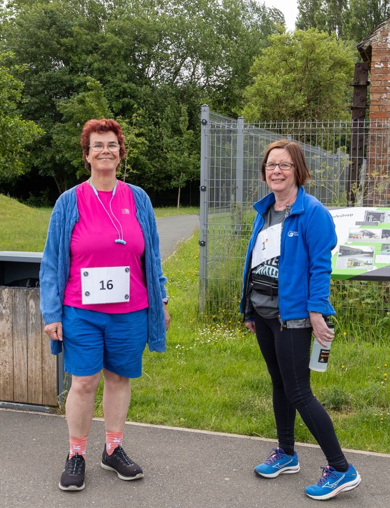 Two runners in athletic wear standing outdoors near a fenced path, one wearing bib number 16.