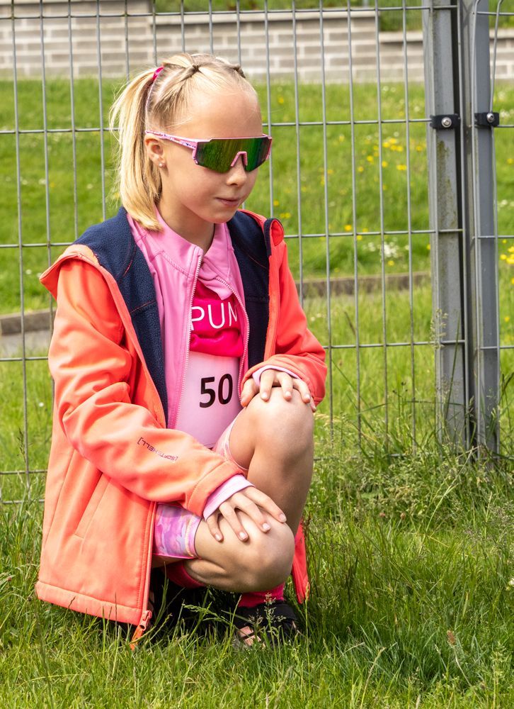 Child sitting on grass by a fence, wearing a coral jacket and pink sunglasses