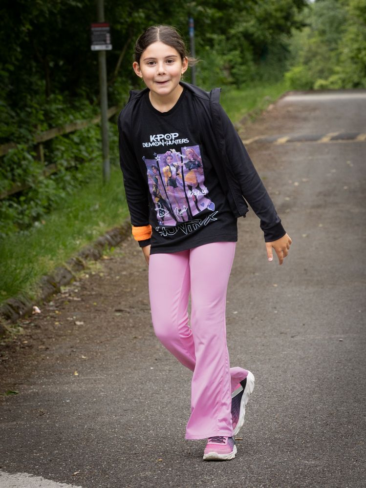 Person walking on a wet road, wearing a black long-sleeve shirt and pink pants, with greenery in the background.