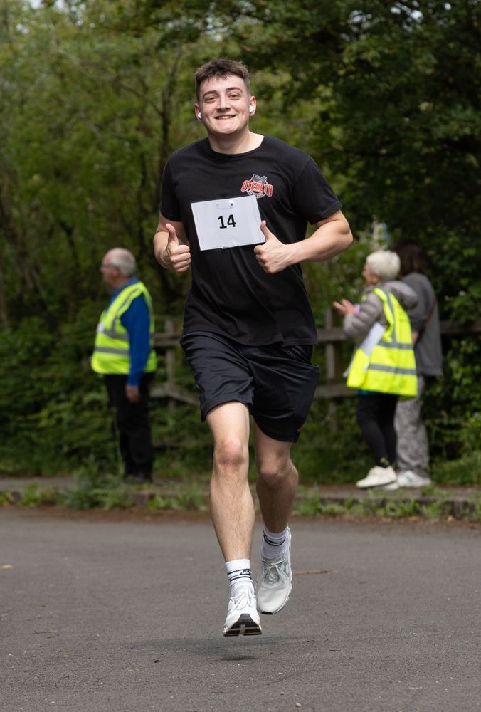 Runner in black outfit wearing bib 14, jogging on a paved path with volunteers in the background.