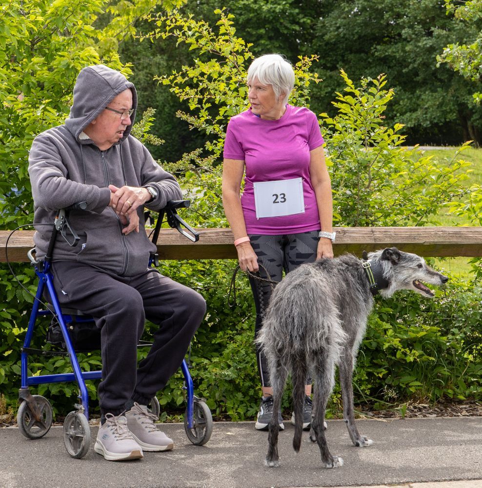 Two people on a sidewalk, one seated on a walker, with a dog and a race bib in a green park area