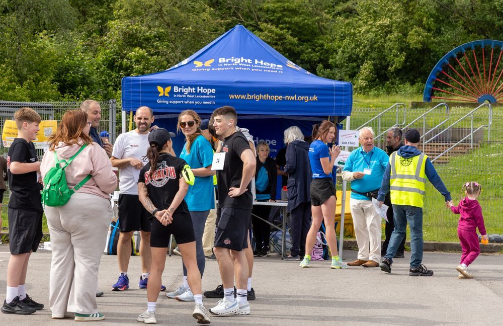 People gathered near a blue charity tent in a park, with volunteers in yellow vests and a Ferris wheel in the background.