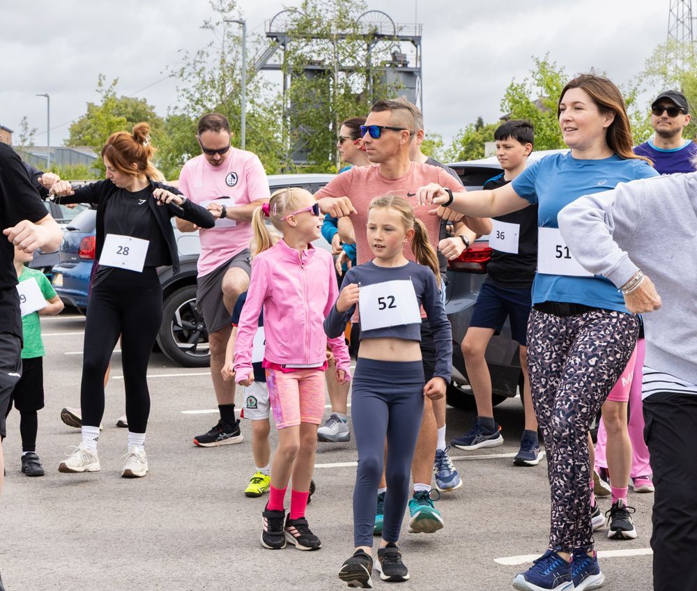Children and adults line up outdoors for a fun run, wearing race bibs and casual athletic clothes.