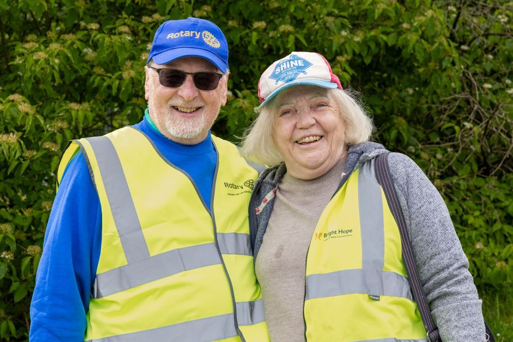Two smiling volunteers in yellow safety vests and blue caps standing outdoors in front of green bushes