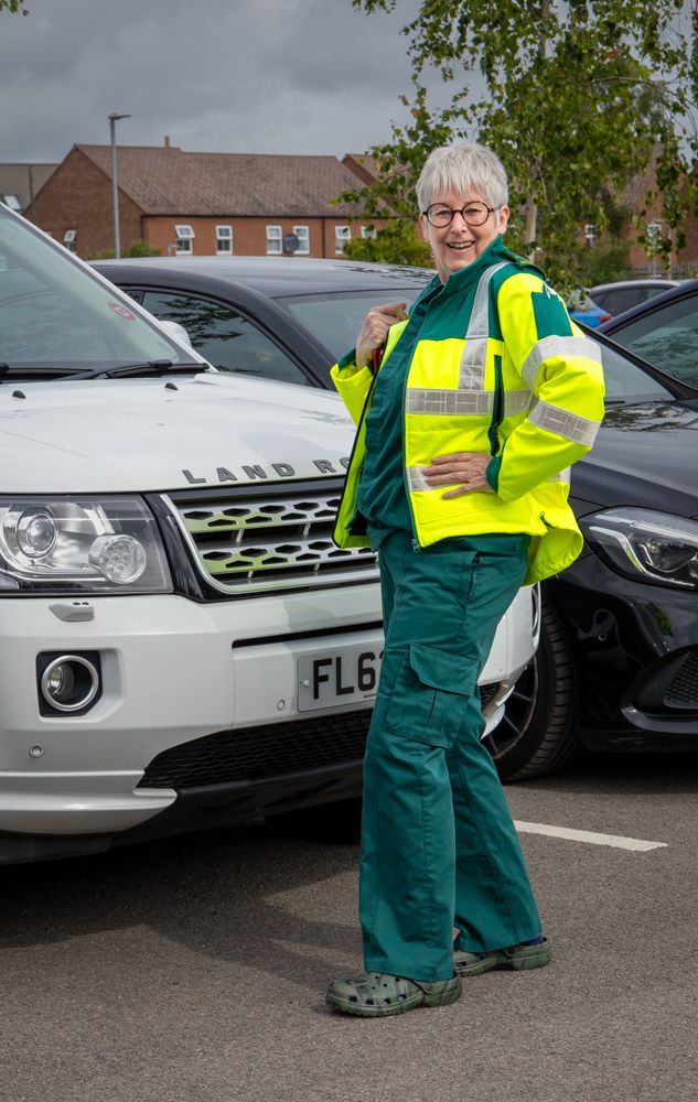 Person in high-vis jacket and green trousers standing in a parking lot beside parked cars.
