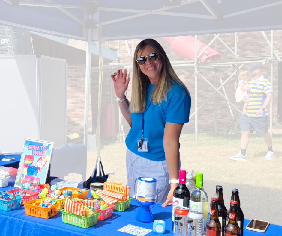 Woman in blue shirt waving at a table of snacks and drinks under a canopy at an outdoor event