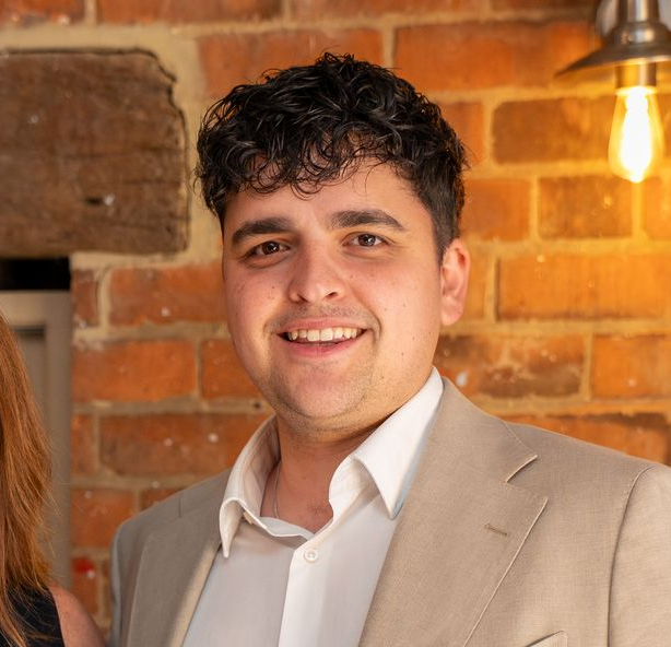 Smiling man in a beige blazer and white shirt standing against a brick wall with warm hanging lights