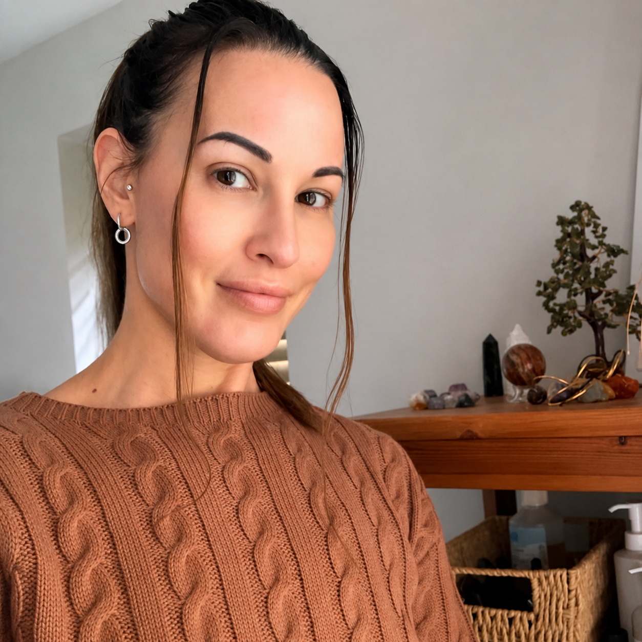 Woman in a brown sweater posing indoors beside a wooden shelf with plants and decor.