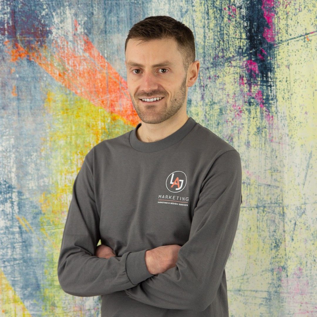 Smiling person with arms crossed in a gray shirt against a colorful abstract wall