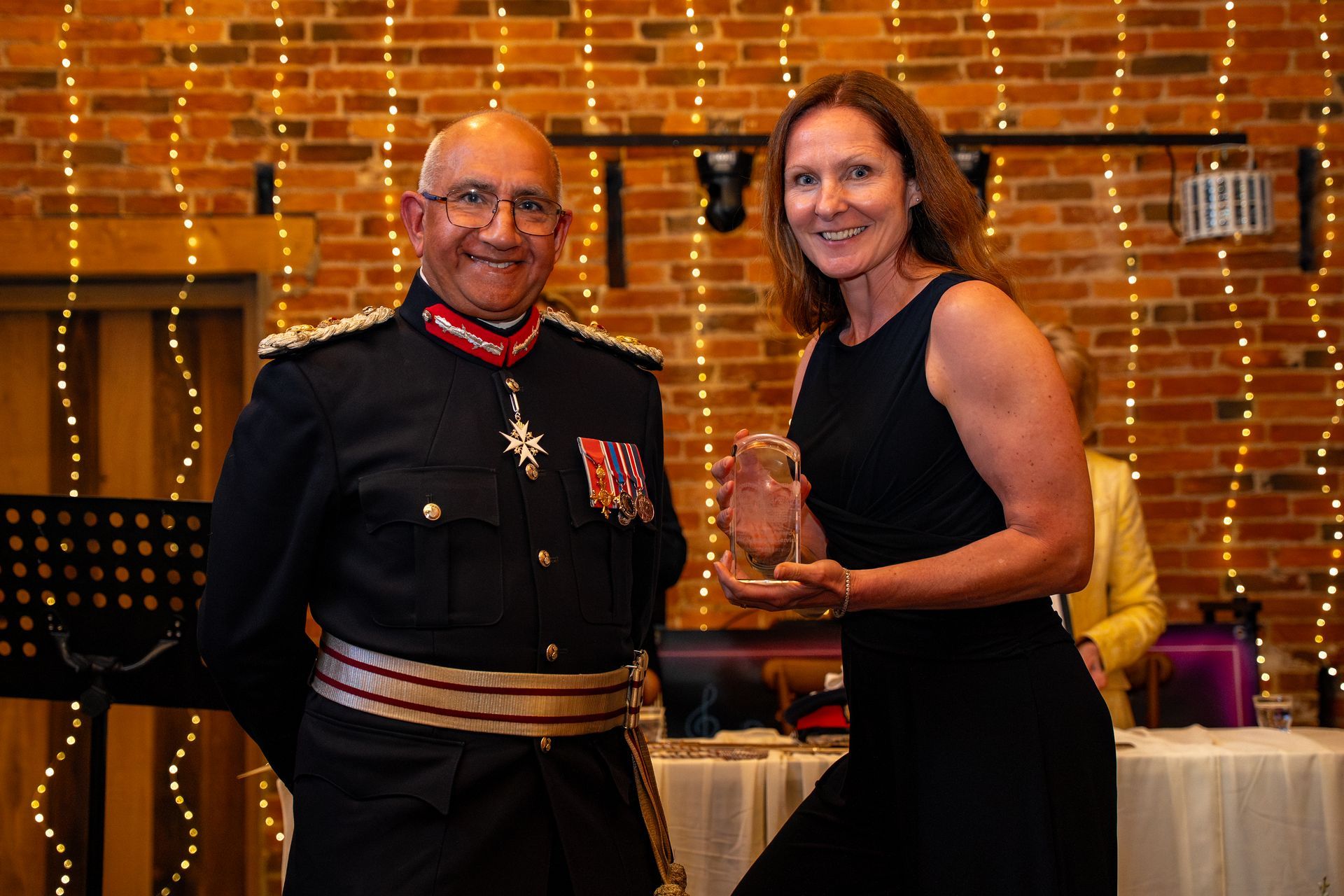 Two people holding an award in a warmly lit event setting with string lights and a table in the background
