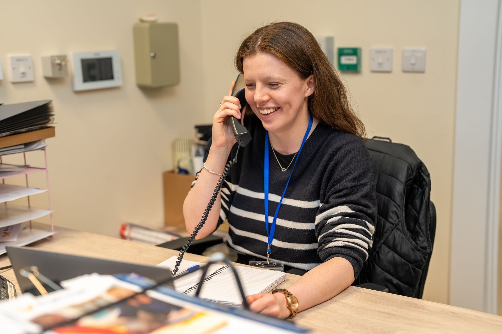 Woman smiling on the phone at a desk in an office, wearing a striped sweater and blue lanyard