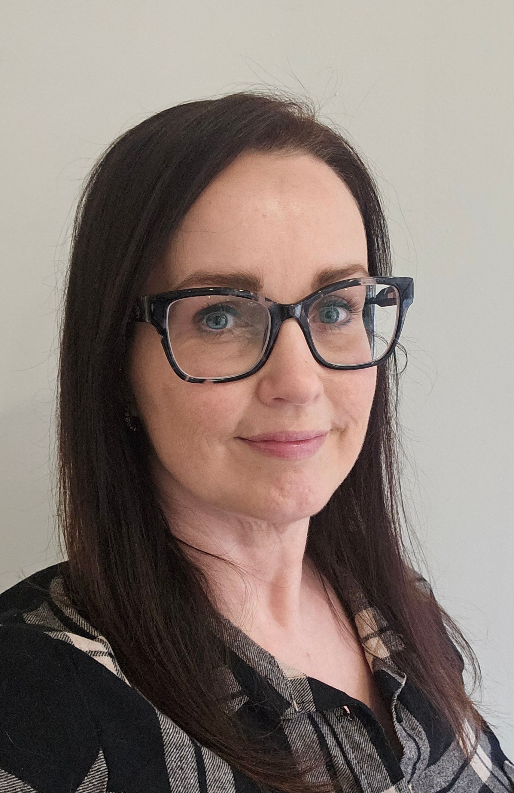 Portrait of a woman with dark hair and glasses, wearing a black patterned top against a plain wall.