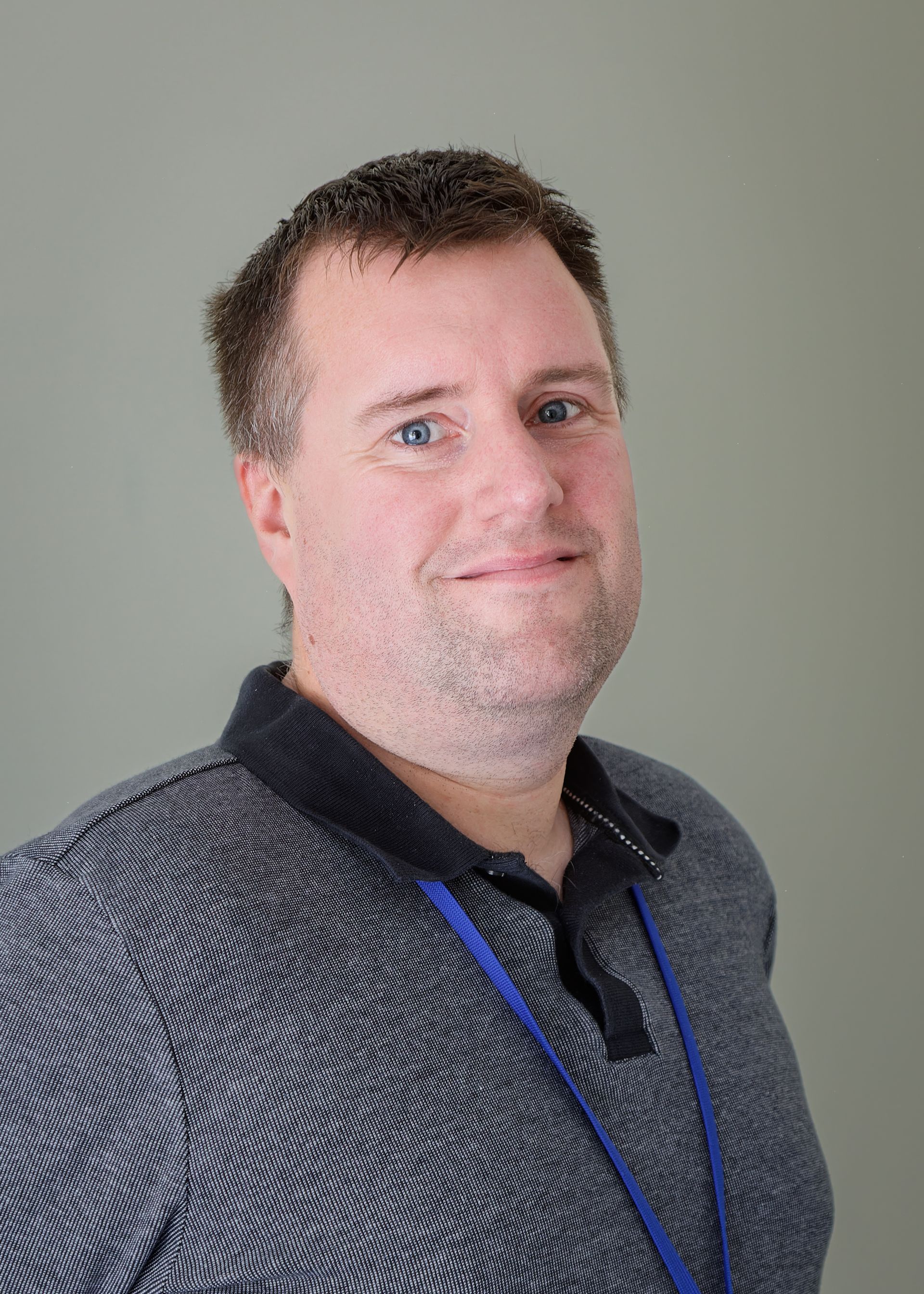 Person in a patterned dark shirt with a blue lanyard, standing against a plain gray background.