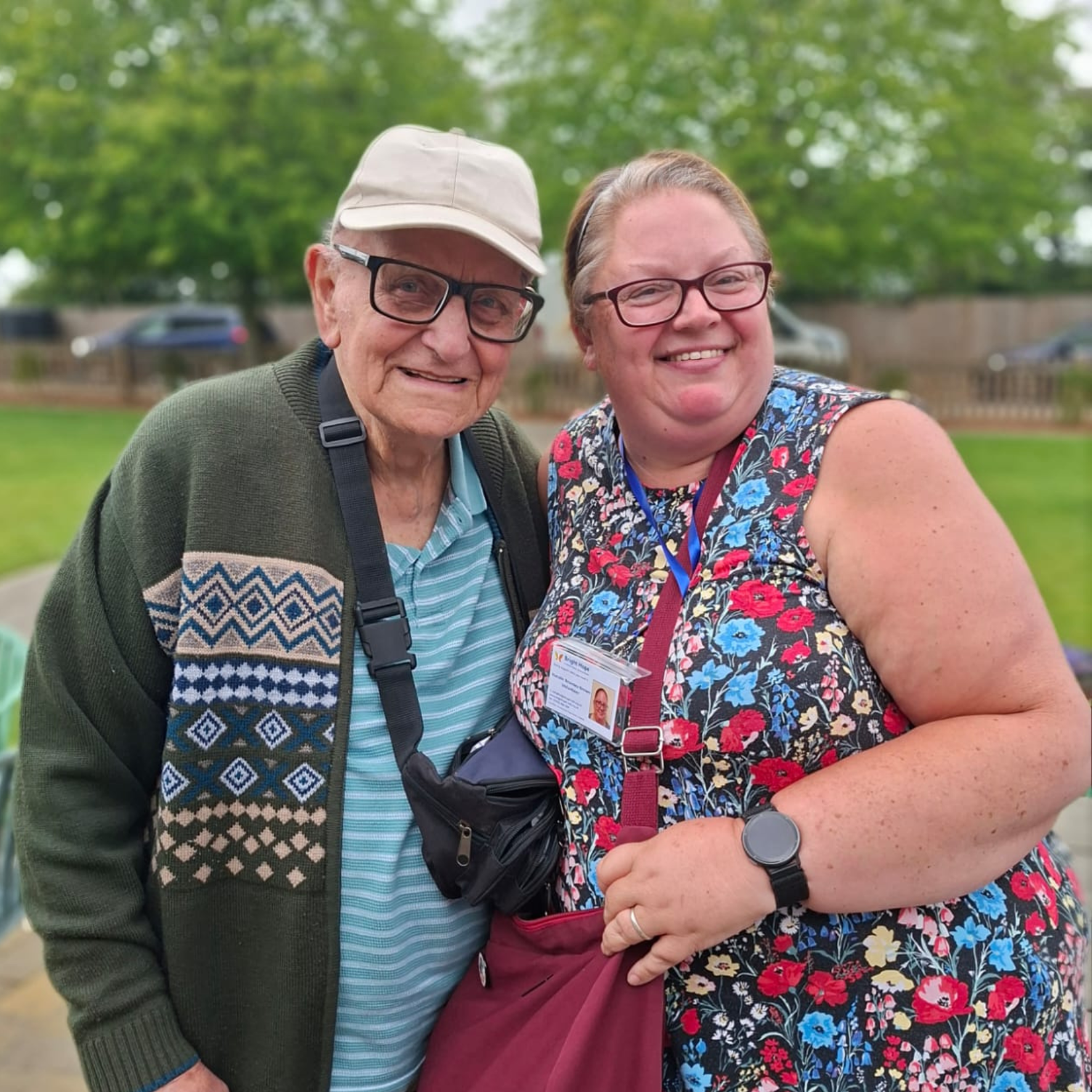 Two smiling people posing outdoors in a park, one in a floral dress and one in a cap and glasses