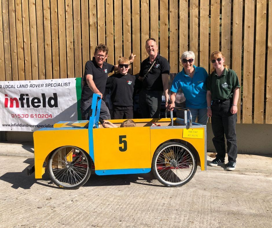Five people stand behind a yellow soapbox car with number 5 in front of a wooden fence.