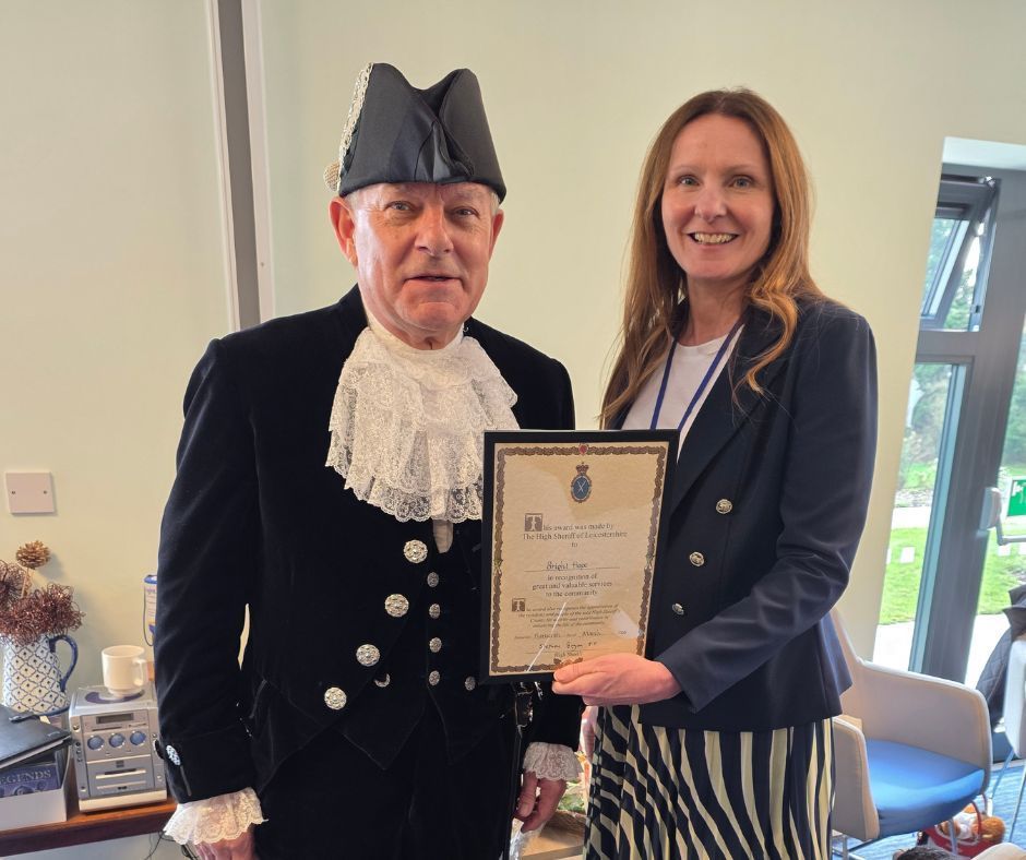 Two people posing with an award certificate indoors, one in formal attire, both smiling.