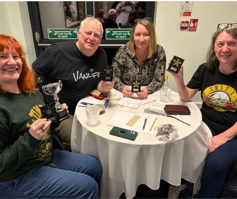 Four people smiling at a round table with papers, pens, and drinks at an indoor event.