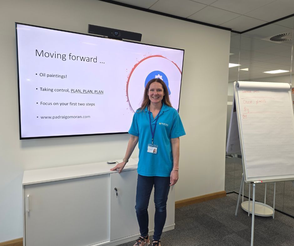 Woman stands beside a presentation screen in an office, smiling and wearing a blue shirt.
