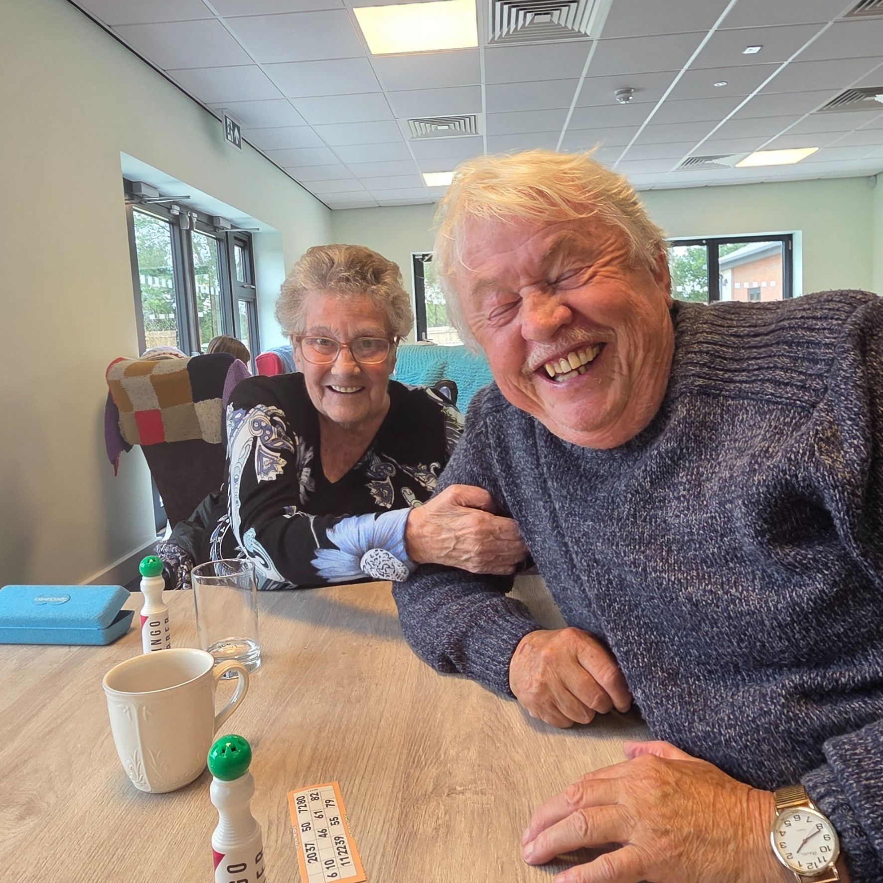 Two smiling people seated at a table in a bright café, leaning toward the camera.
