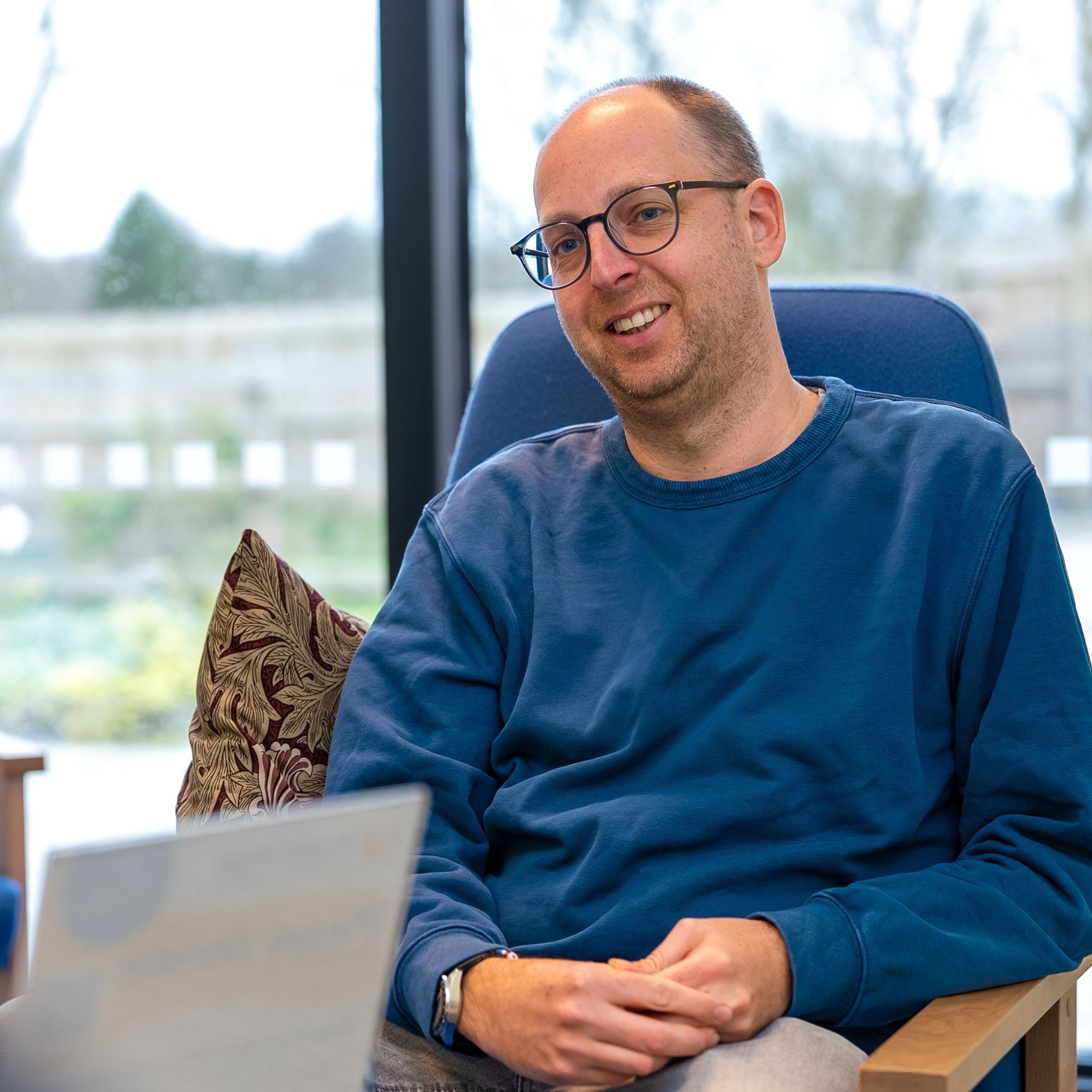 Man in glasses smiling in a blue sweater, seated indoors by a bright window with a laptop in the foreground