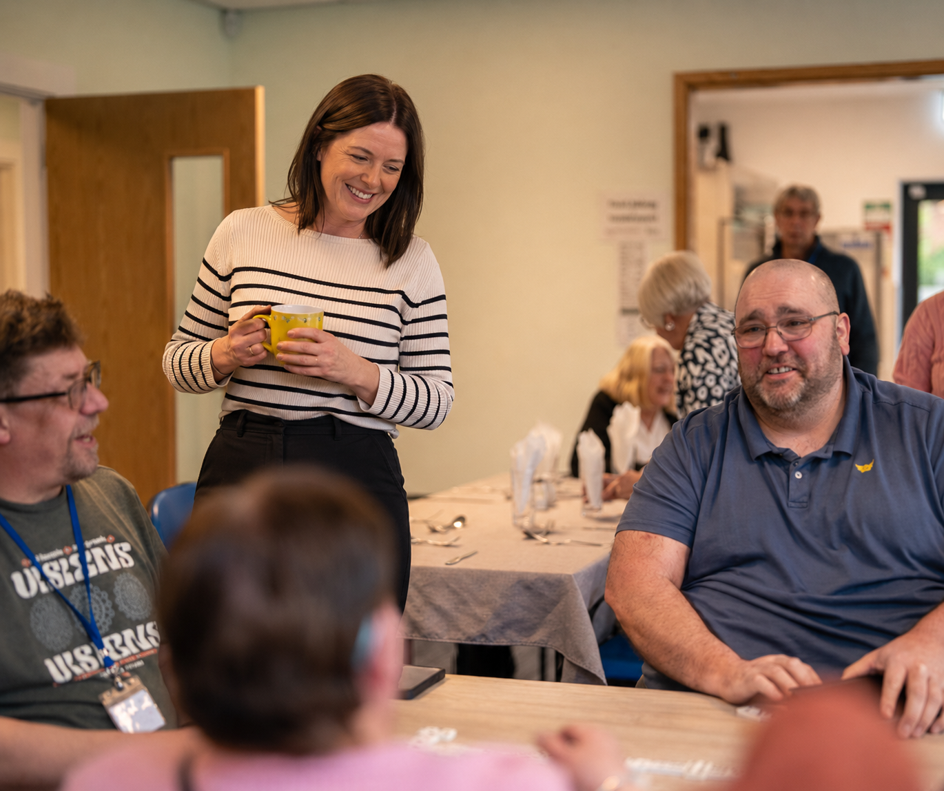 People gathered in a meeting room; a woman stands smiling and speaking beside seated attendees.
